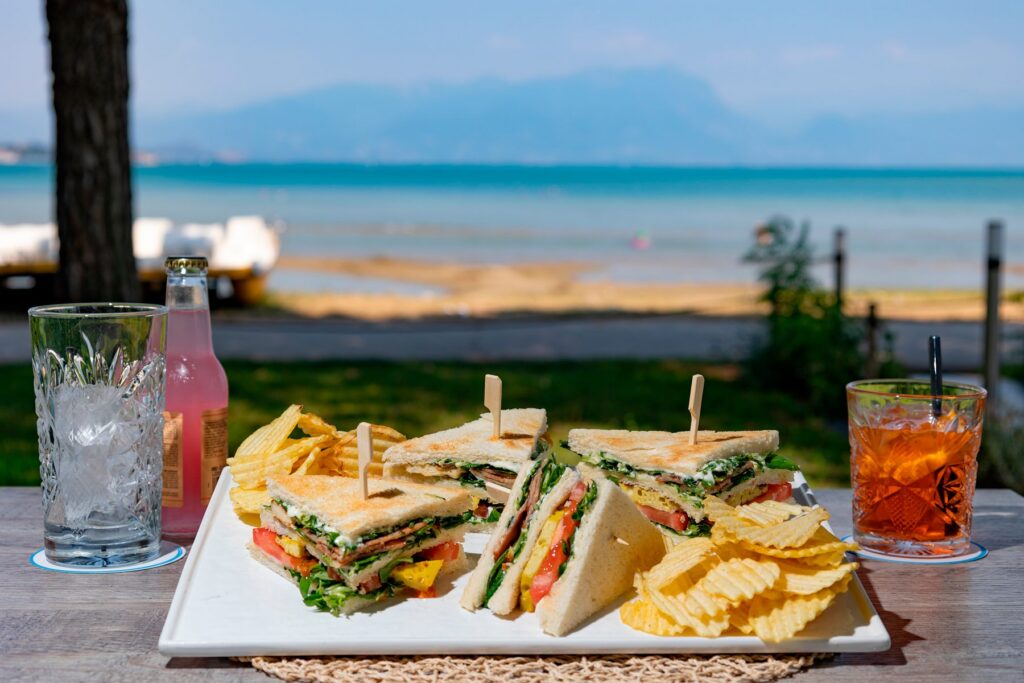 Club sandwich, patatine e spritz sul tavolo, con il lago di Garda sullo sfondo
